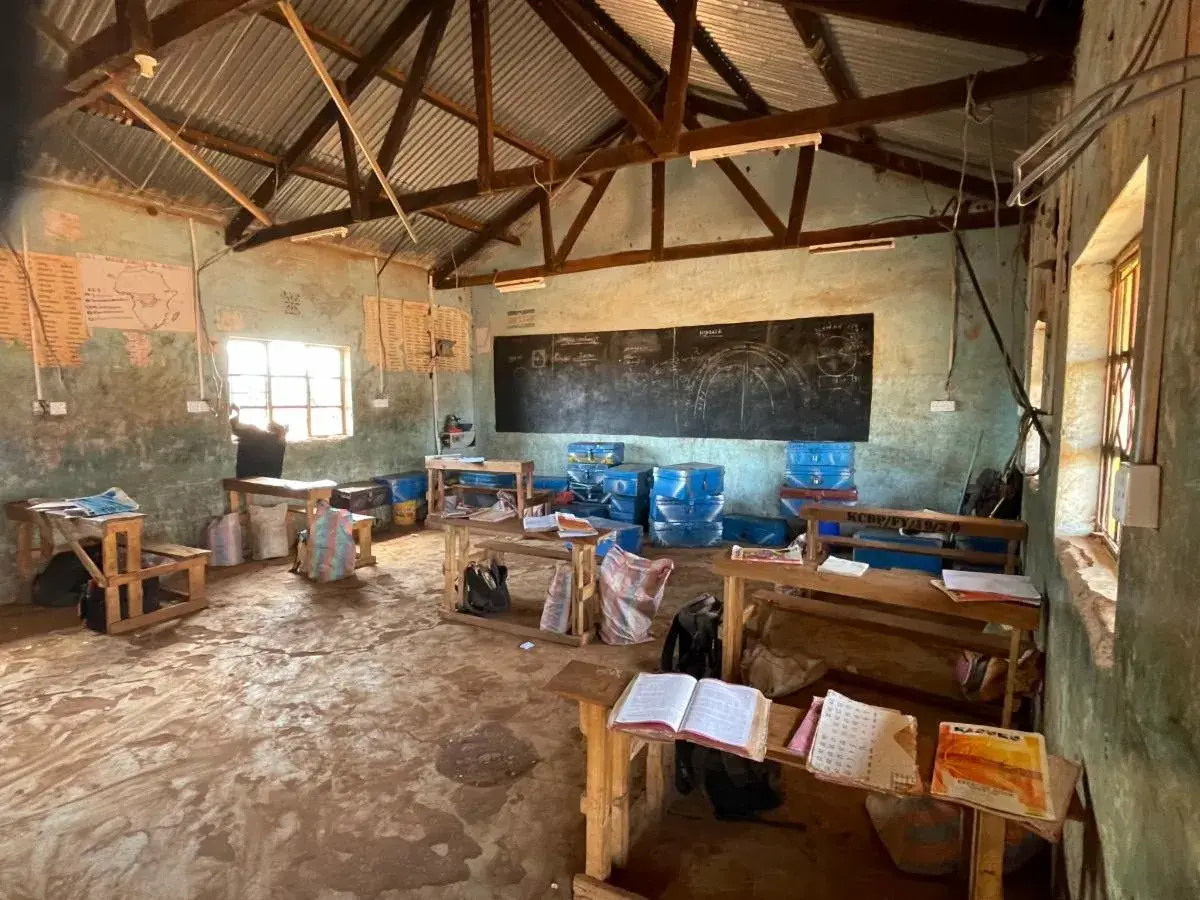Inside a rural classroom with a corrugated iron roof: wooden desks and benches on a bare earth floor, a blackboard on the wall and open books lying on the desks