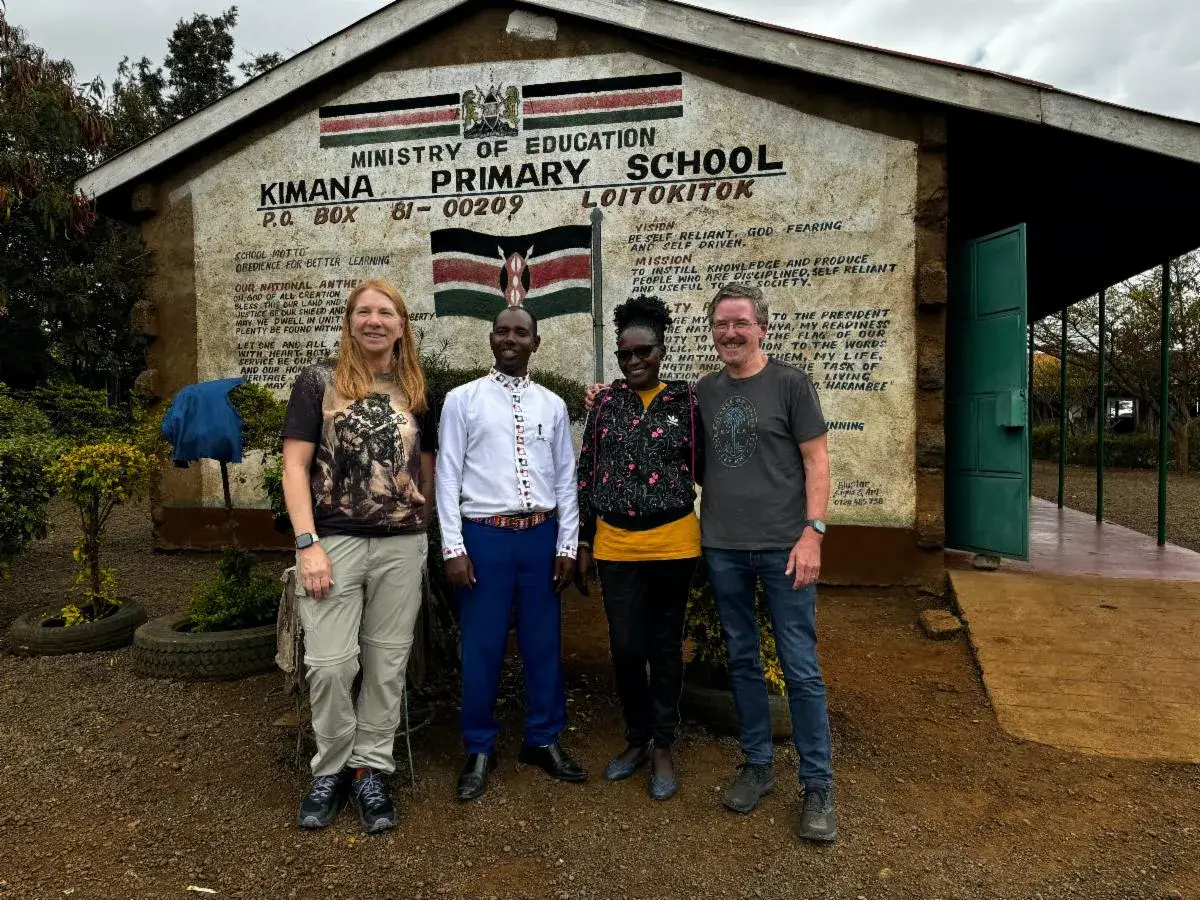 Four adults stand in a row in front of the painted sign of Kimana Primary School: a woman with long fair hair, a man in a white shirt and blue trousers, a woman in a black top and yellow skirt, and a man in a grey t-shirt