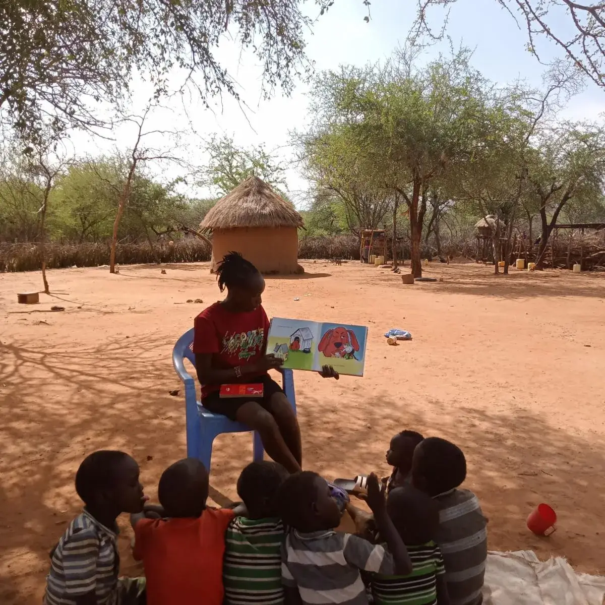 A girl in a red top sits on a blue plastic chair reading a picture book aloud to five children sitting on the red earth in front of her, under acacia trees beside a thatched hut