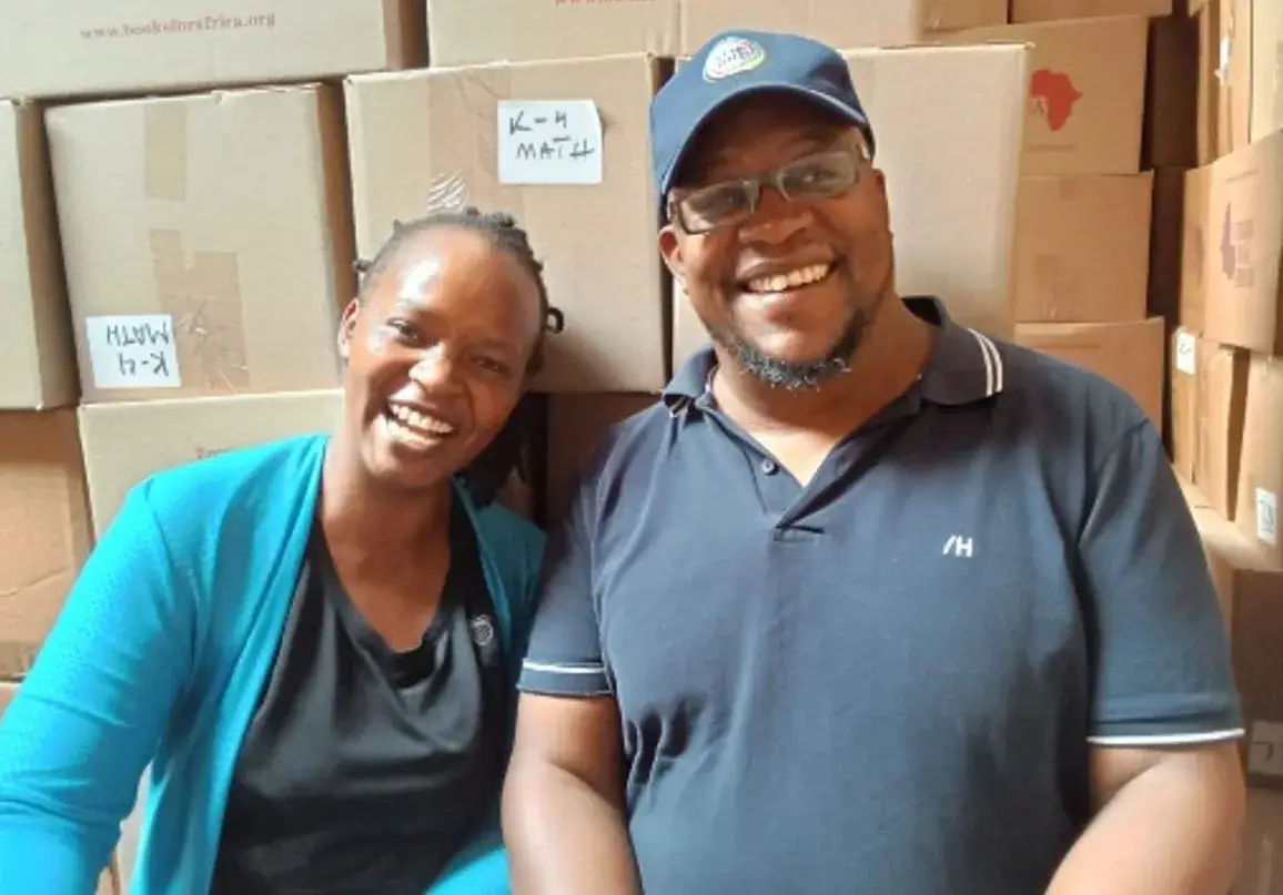 A woman in a teal jacket and a man in a blue polo shirt smiling side by side in front of stacked cardboard boxes