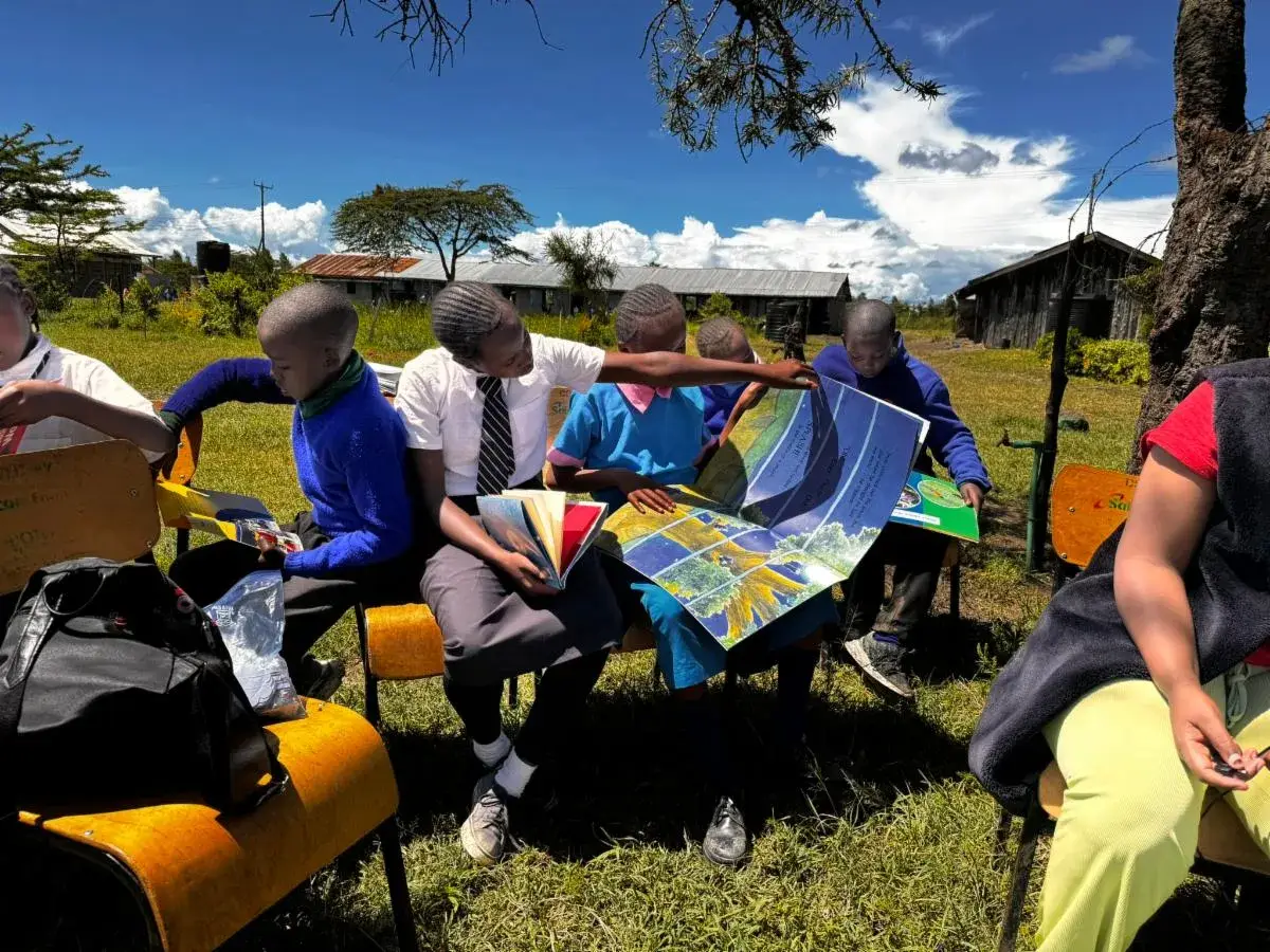 Students in school uniform sit outdoors on yellow chairs with a large colourful map spread across their laps, school buildings behind them