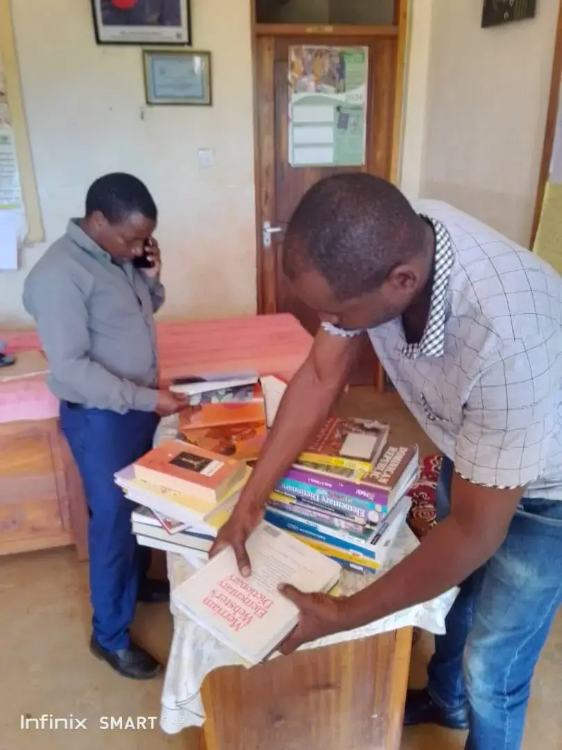 The headteacher and a teacher of Kisiwani Primary School in a school office, one speaking on the phone and the other bending over a desk to look through a pile of books