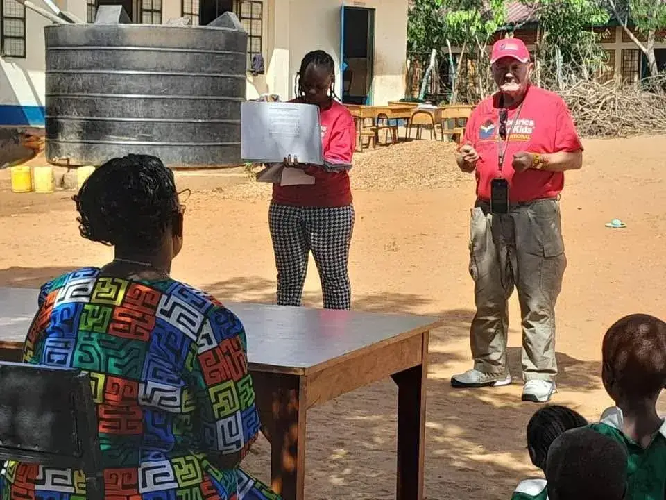 A man in a red cap and red shirt speaks to a small group outside a school building with a water tank behind it, a woman holding up a sheet of paper beside him, and a woman in a bright patterned dress and pupils in the foreground