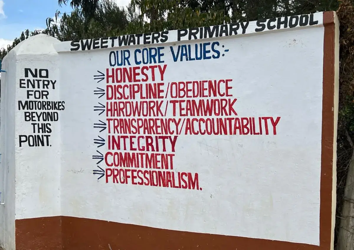 The painted wall sign of Sweetwaters Primary School listing its core values: Honesty, Discipline/Obedience, Hardwork/Teamwork, Transparency/Accountability, Integrity, Commitment and Professionalism, with a notice that motorbikes may not go beyond this point