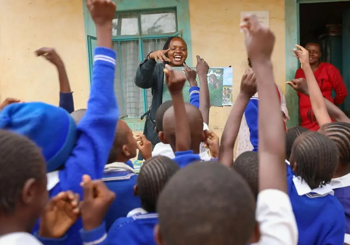 Pupils in blue jumpers sitting on the floor of a classroom with their hands raised, a teacher standing at the back