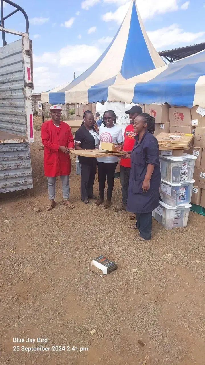 A group of about six people in red and dark clothes stand on red earth in front of a blue and white striped tent beside a truck, with stacked plastic crates and boxes at their side, as the Grand Circle Foundation container is loaded