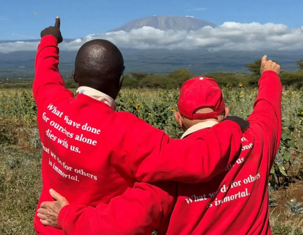 Two men in red jackets seen from behind, arms around each other, pointing at the snow-capped top of Mount Kilimanjaro above farmland