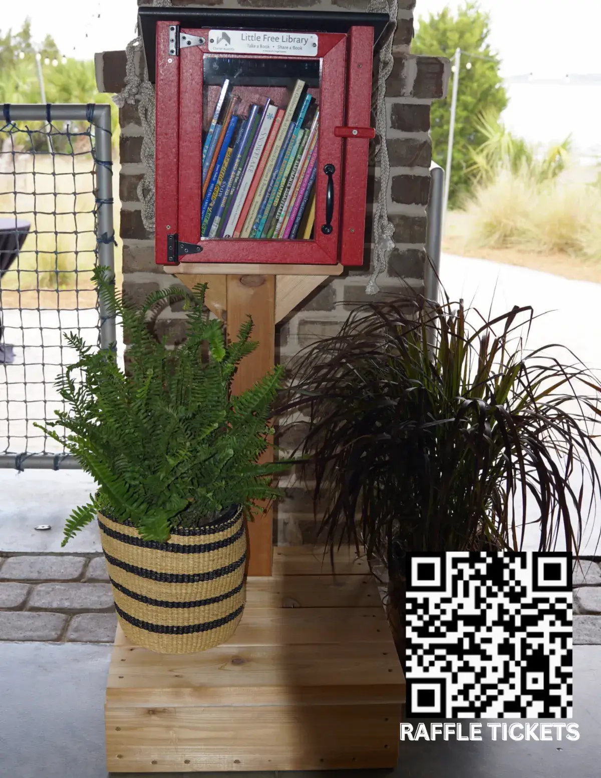 A red Little Free Library box with a glass door, filled with books, mounted on a post beside a potted plant and a basket, with a QR code labelled Raffle Tickets in the corner