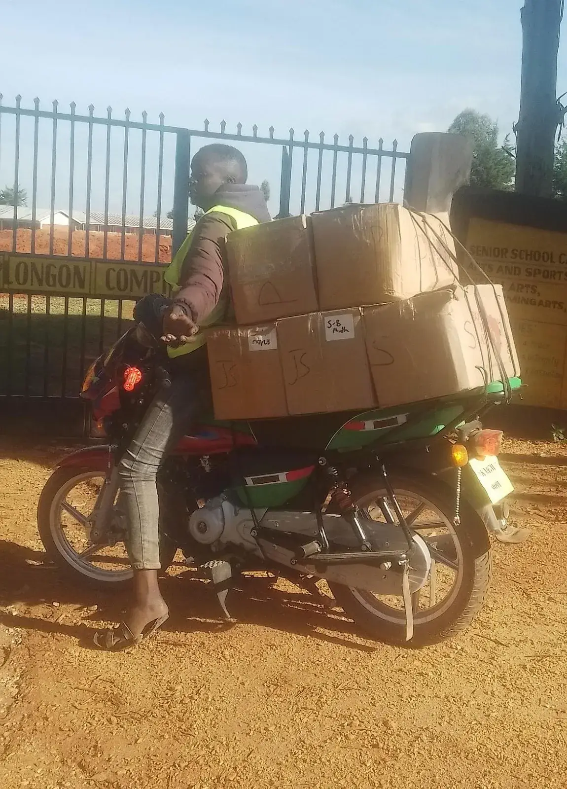 A man in a high-visibility vest riding a motorbike loaded high with cardboard boxes, on red earth beside a fence