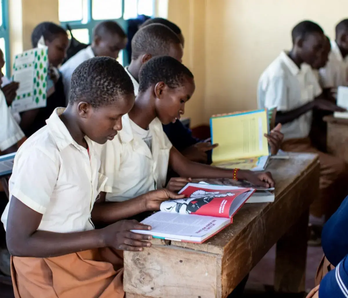 Pupils in white shirts sharing open books at a wooden desk in a classroom, with more pupils at desks behind them