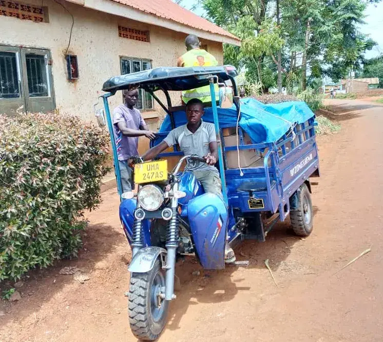 A blue three-wheeled cargo vehicle with a driver and passengers on a dirt road in front of a school building in Uganda