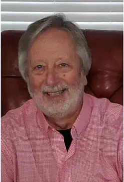 William Douglas, the author, smiling, with grey hair and beard and a pink shirt, seated in a leather chair
