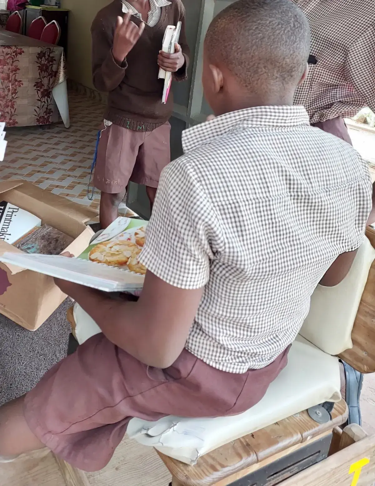 A boy in a checked shirt and shorts seen from behind, seated on a chair and looking into an open box of books