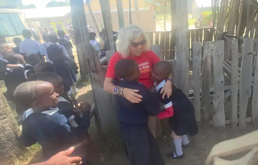 A woman in a red T-shirt crouching to hug a pupil in a school uniform outdoors, with other pupils in dark uniforms sitting and standing around them