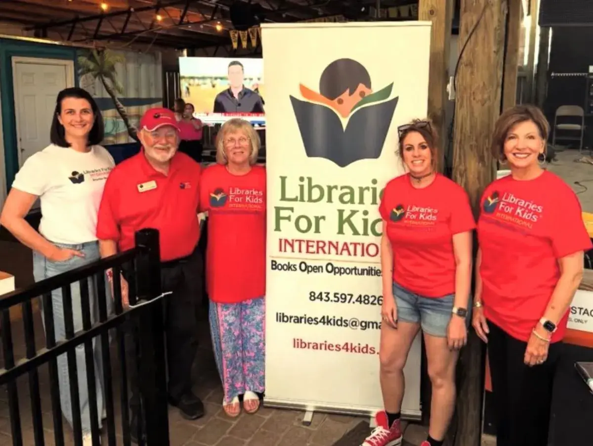 Six Libraries For Kids International volunteers, most in red shirts, standing in front of a pull-up banner at an indoor venue