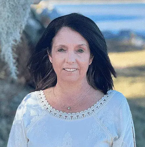 Laura Elizabeth, the author, smiling, with shoulder-length dark hair and a white lace-trimmed top, with the sea behind her