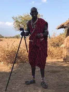 A Maasai man in a red checked shuka standing beside a camera on a tripod on dry ground