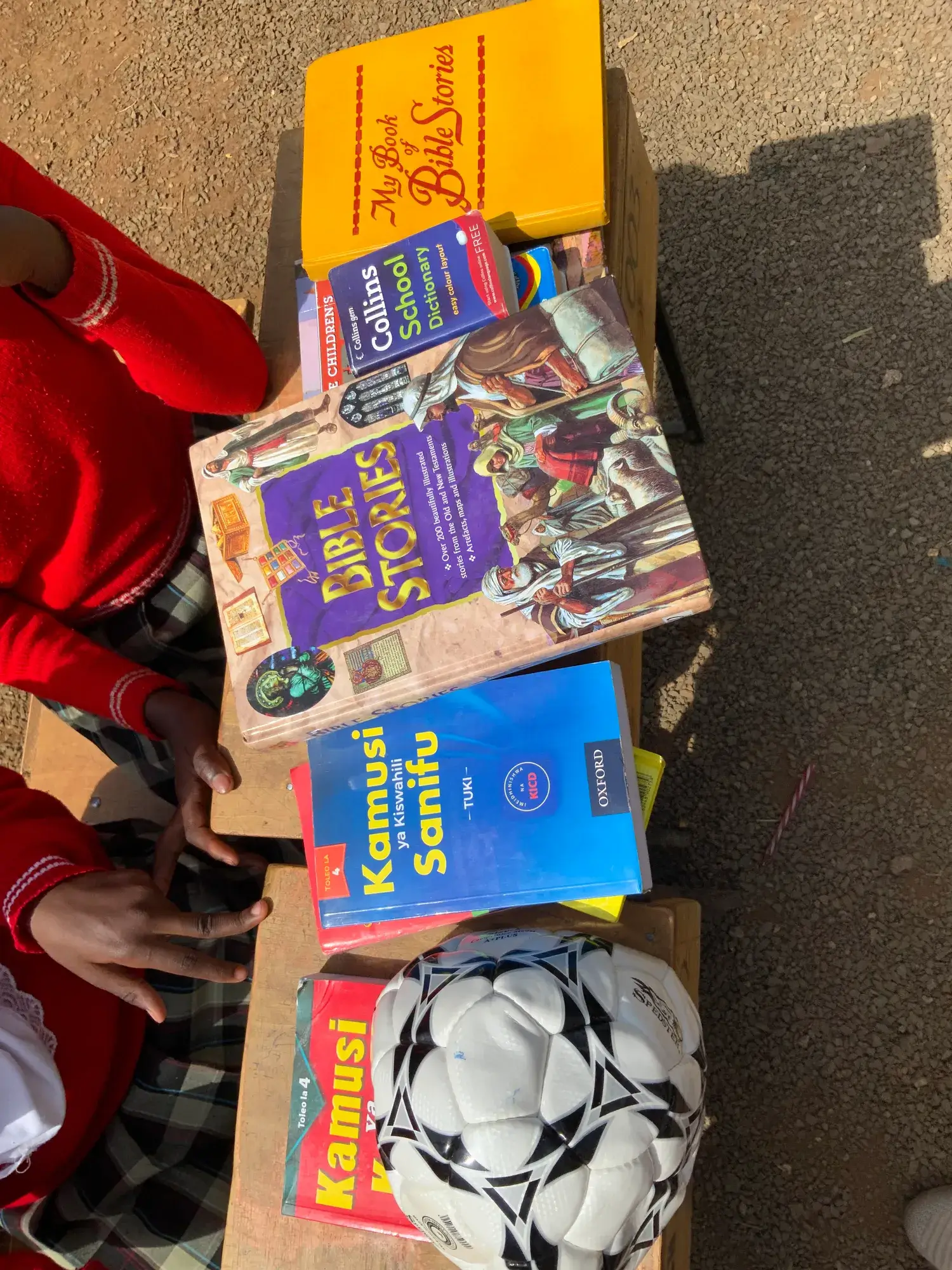 Bible story books, a school dictionary and Swahili dictionaries on a desk beside a football, pupils in red sweaters alongside