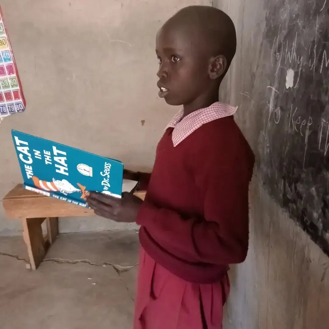 A student in a maroon sweater holding The Cat in the Hat beside a classroom chalkboard