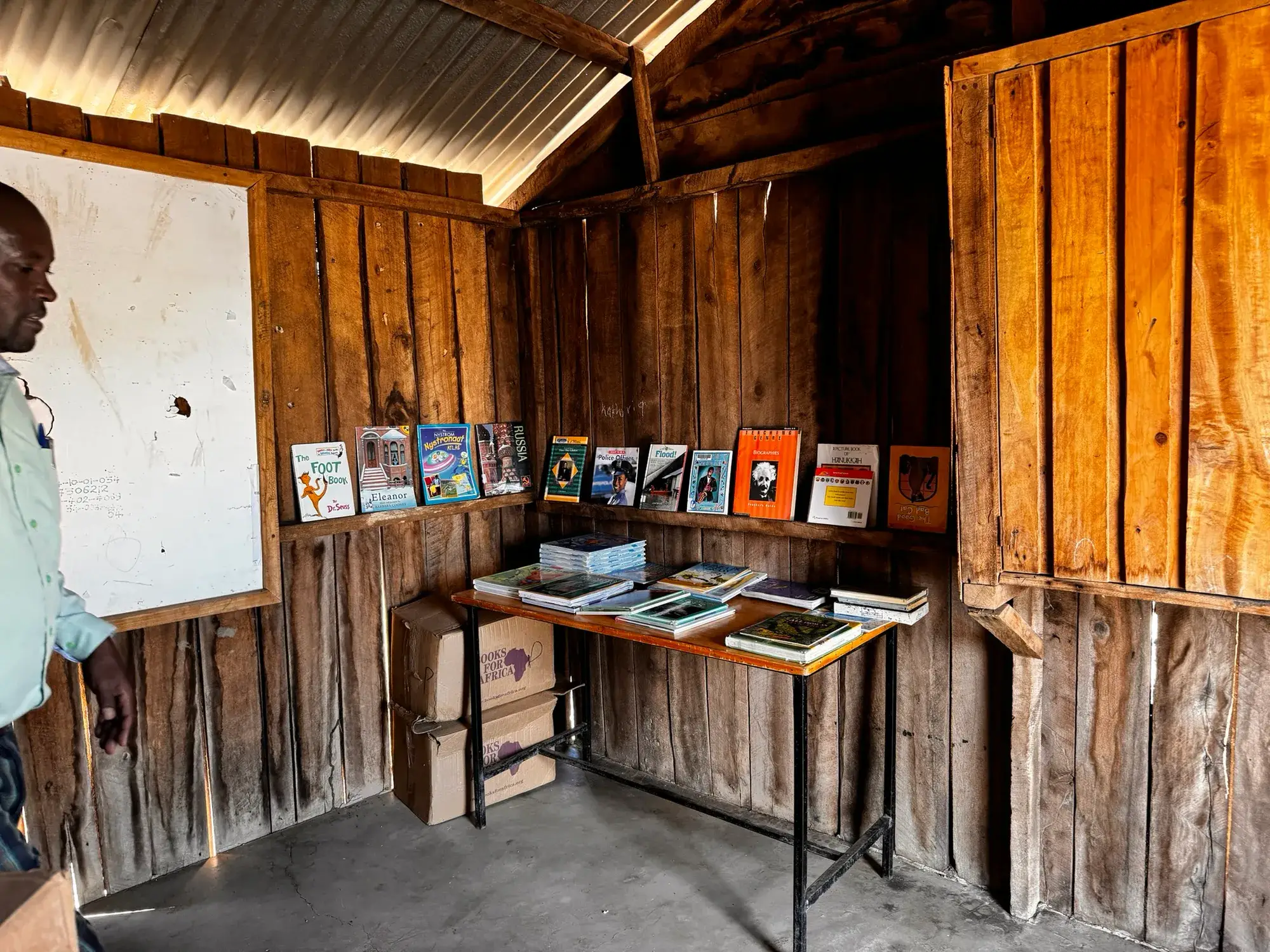 A small school library with plank walls and a tin roof, picture books standing along a ledge and laid out on a table, a teacher by the whiteboard