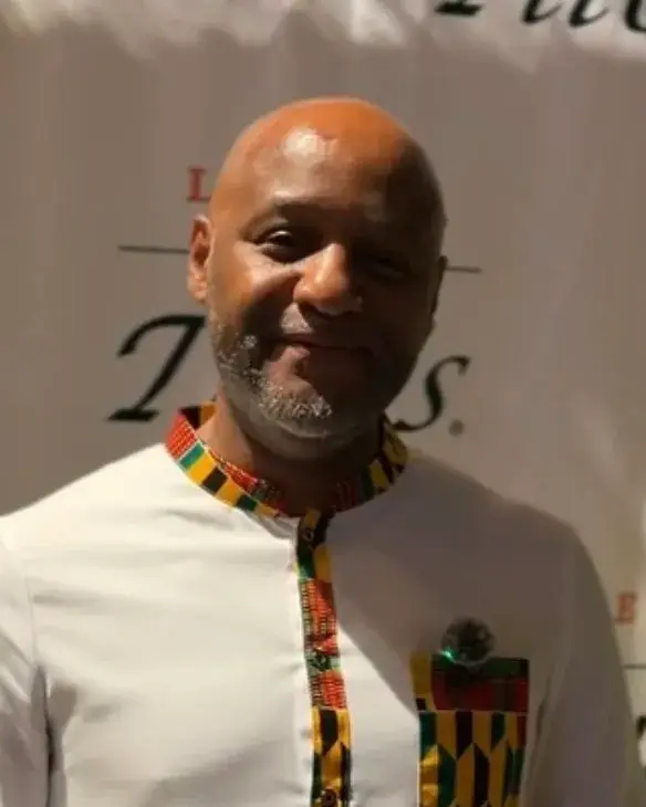 A smiling man with a shaved head and a grey beard, in a white tunic with a bright woven-pattern collar
